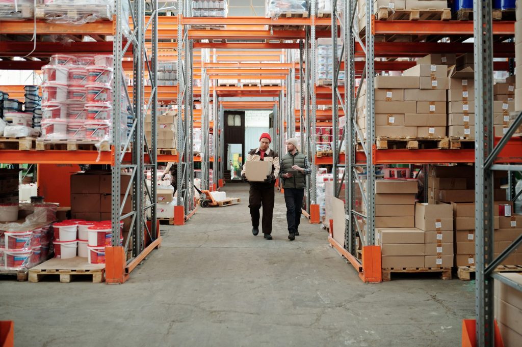 Two logistics workers walking through a warehouse aisle, one carrying a parcel and the other checking a tablet